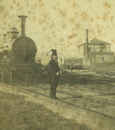 Sepia photograph of a man dressed in a top hat and frock coat standing before a steam engine on train tracks. There are sheds and buildings in the background and some age or water damage to the photograph.